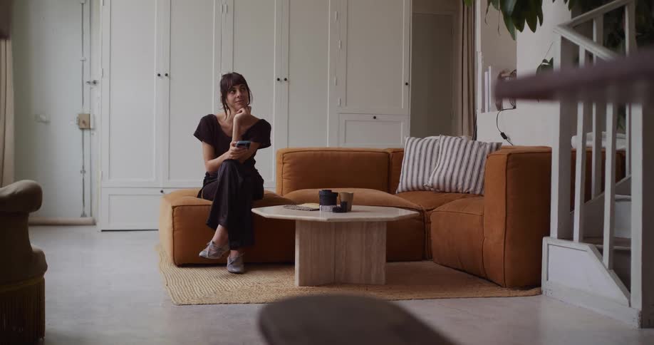 A thoughtful woman relaxing on a stylish brown sofa scrolls on her phone in a modern living room