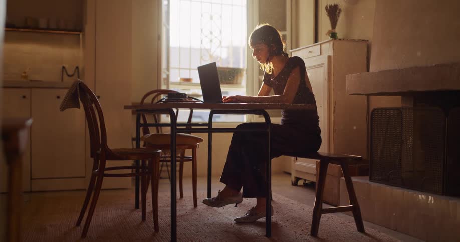 A woman works on her laptop at a table in a dimly lit rustic room, with warm sunlight and dust particles creating a cinematic atmosphere