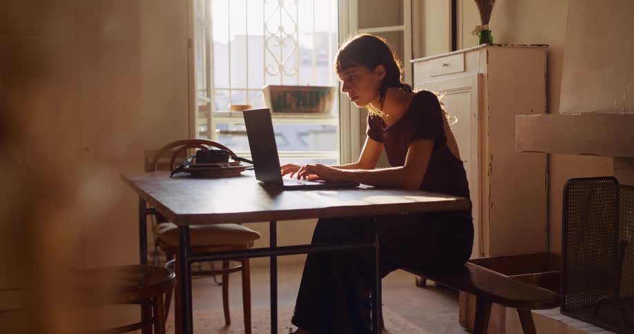 A woman works diligently on her laptop at a wooden table in her cozy, sunlit apartment