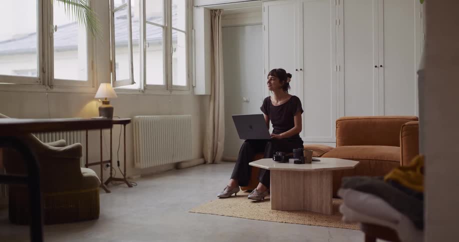 A thoughtful woman works on her laptop in a chic, naturally lit studio apartment