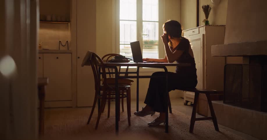 A woman works on her laptop at a dining table in a cozy, sunlit apartment and then takes a phone call