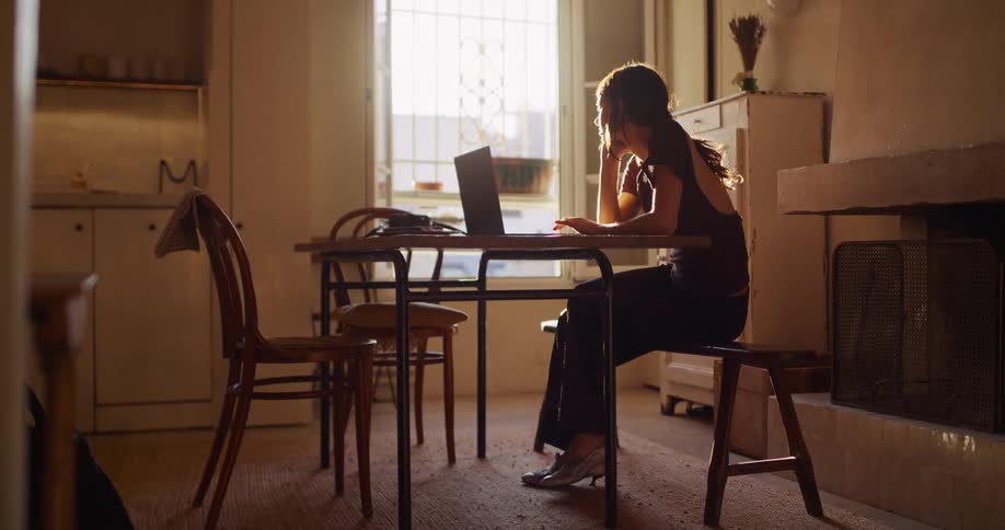 A focused woman works on her laptop at a dining table, beautifully backlit by the warm, natural light from a window