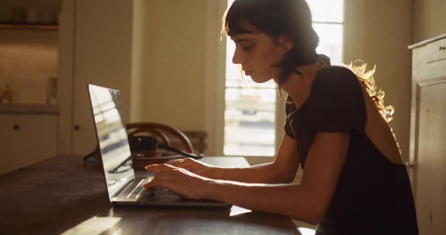 A focused woman works on her laptop at a wooden table, bathed in the warm, dusty sunlight streaming through a window in her home
