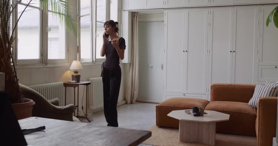 A stylish woman has an engaging phone conversation while walking around her modern, sunlit apartment