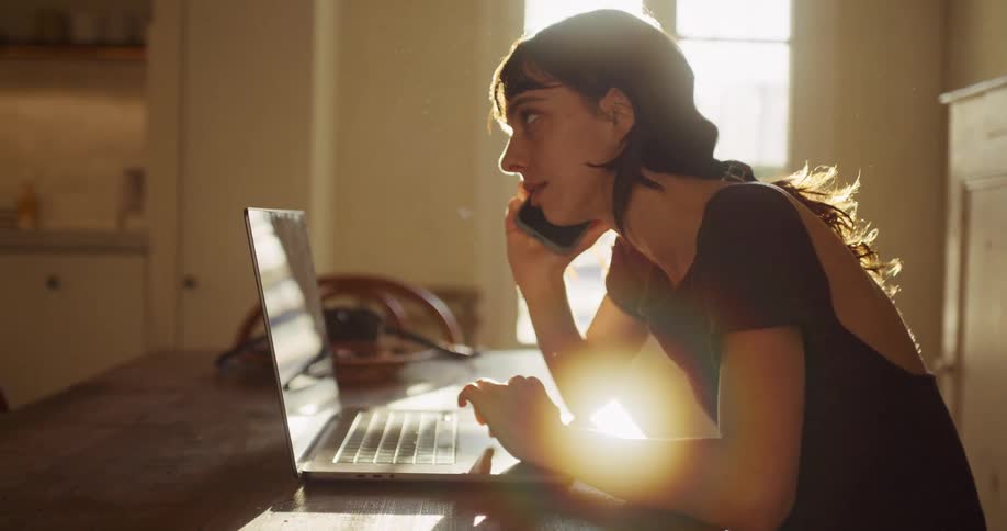 A woman sits at a wooden table in a sun-drenched room, smiling while talking on the phone and looking at her laptop