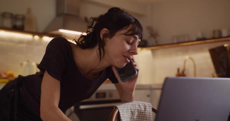 A happy young woman multitasks, talking on her phone while looking at her laptop in a modern, warmly lit kitchen