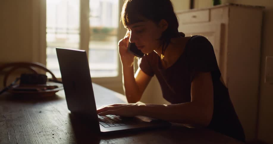 A woman multitasks on her phone and laptop, sitting at a table bathed in warm, natural light from a window