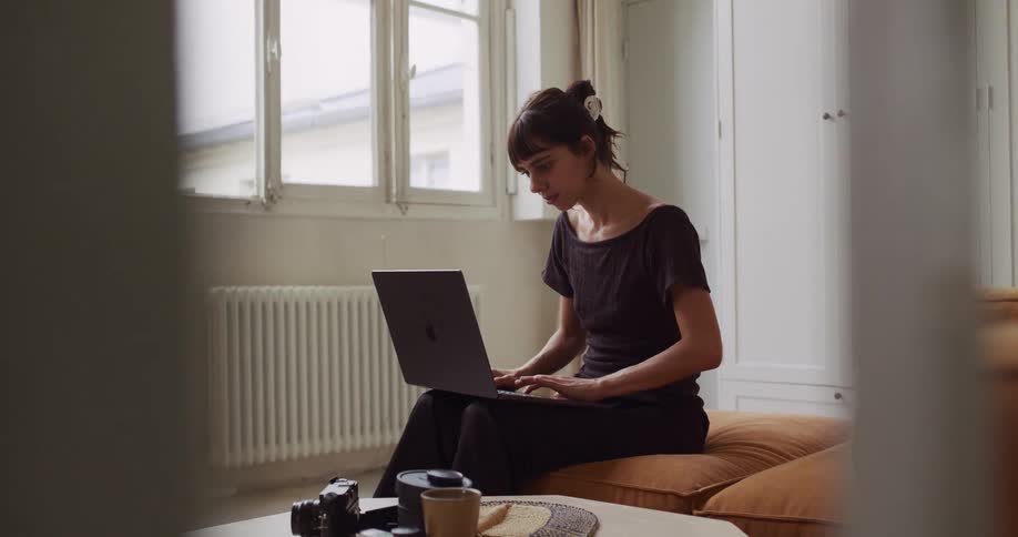 A young woman working from home on a couch closes her laptop and pauses to look thoughtfully out the window