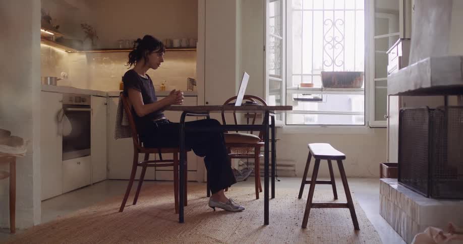 A woman takes a brief stretch break while working on her laptop in her cozy, sunlit apartment kitchen