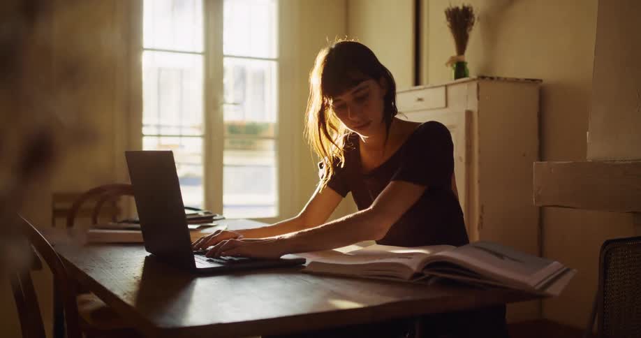 A focused young woman researches in a book before typing on her laptop while studying in her sun-drenched apartment