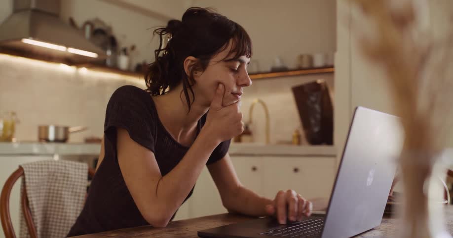 A young woman works diligently on her laptop at a wooden table in her cozy, sunlit kitchen