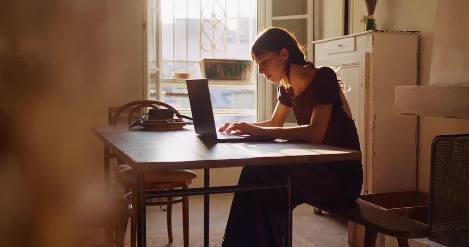 A young woman sits at a rustic wooden table and works on her laptop as the warm sunlight streams into her apartment