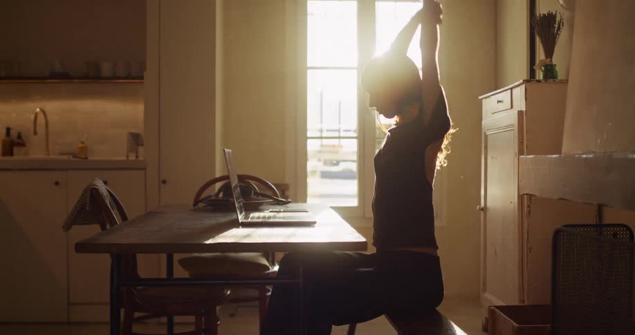 A silhouetted woman works on her laptop in a sunlit room, pausing to stretch her arms and tie up her hair