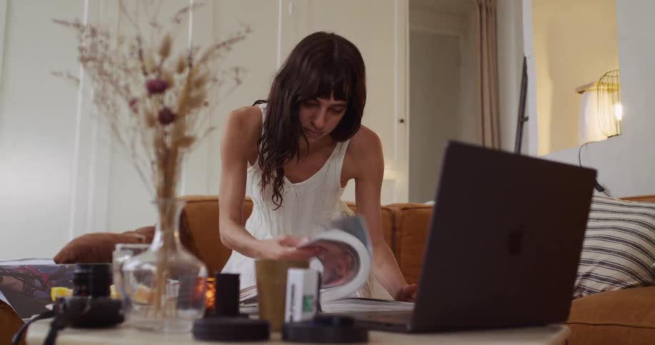A focused woman in a white dress flips through a book for inspiration in her cozy living room