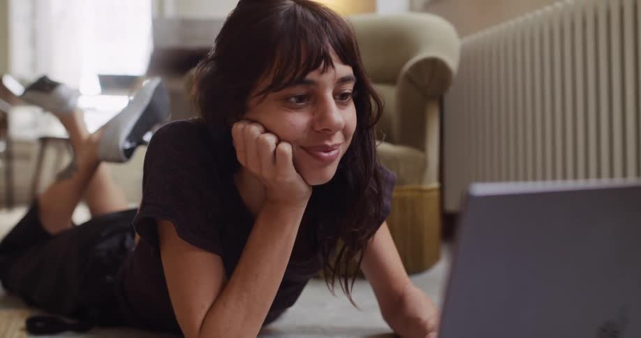 A young woman with a pleasant smile lies comfortably on the floor of her home while using a laptop computer