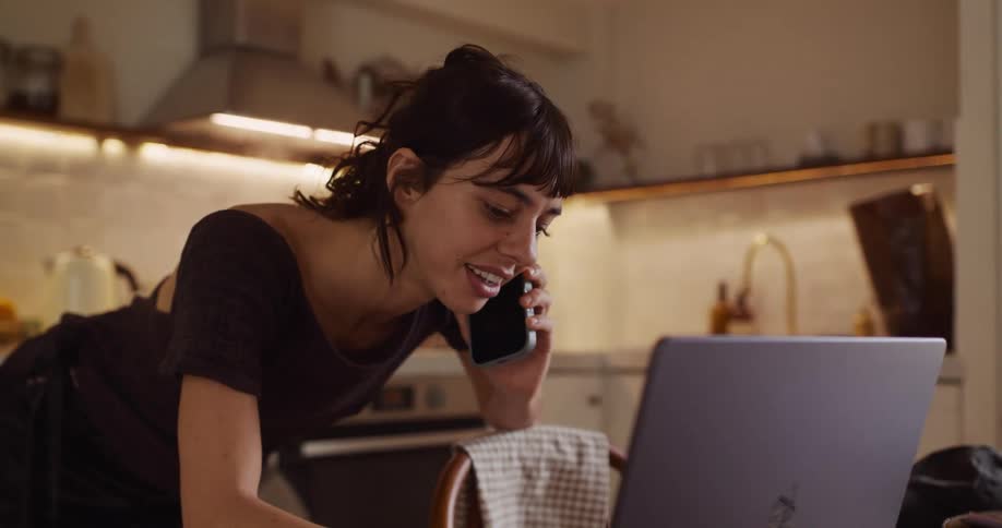 A smiling young woman multitasks by talking on her phone and working on her laptop in her cozy home kitchen