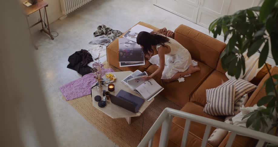 A creative woman in a white dress gathers inspiration by flipping through art books while working on her laptop from her cozy living room couch