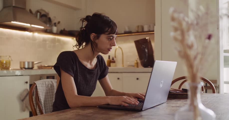 A focused young woman works remotely on her laptop at a wooden table in her cozy, sunlit kitchen