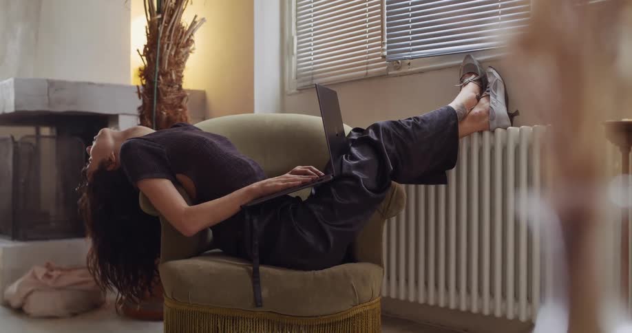 A young woman comfortably works on her laptop in an armchair at home, stretching back tiredly before returning to her work