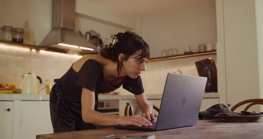 A young woman stops cooking to urgently work on her laptop in the kitchen
