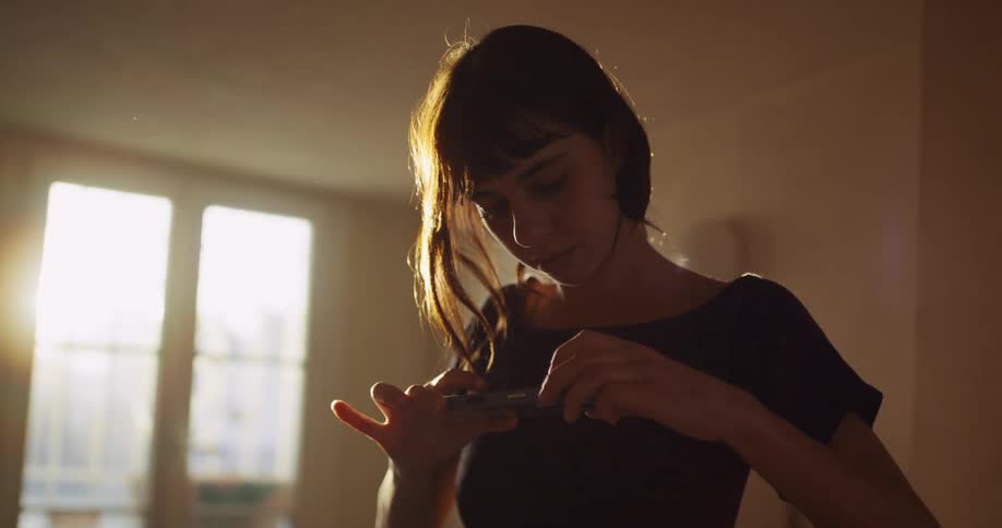 A focused young woman stands in a dimly lit room, her face illuminated by her smartphone as strong sunlight streams in through a window behind her