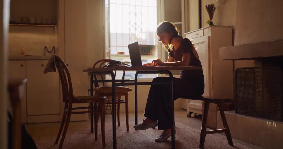 A focused woman works on her laptop at a dining table in a rustic, sunlit room