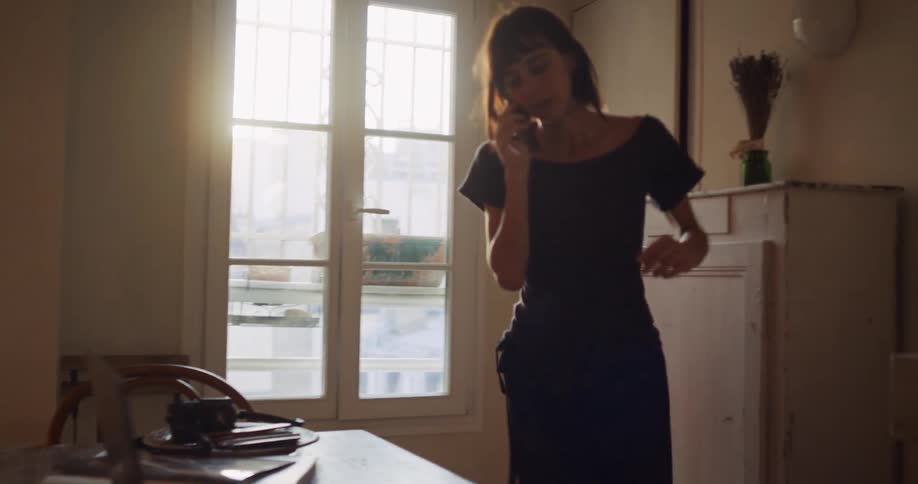 A woman stands by a sunny window talking on the phone before moving to her desk to work on her laptop in a chic apartment