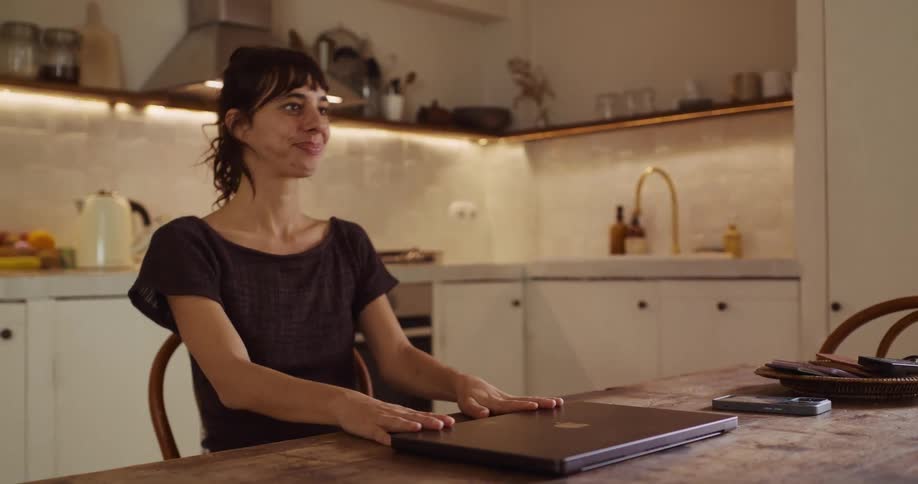 A smiling woman finishes her remote work for the day, closing her laptop with satisfaction in a cozy kitchen