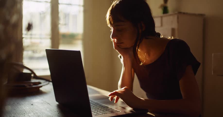 A focused young woman works on her laptop at a rustic wooden table as golden hour sunlight streams through the window