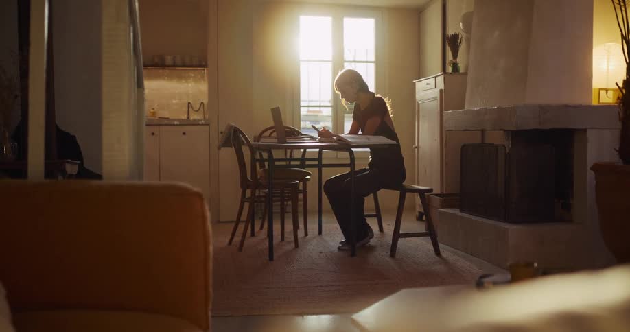 Backlit by the warm sun, a woman sits at her table with her laptop and scrolls on her phone in her cozy apartment
