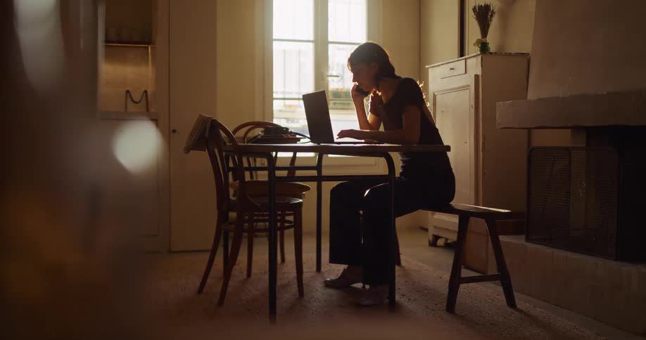 A young woman multitasks on her laptop and a phone while working from home in a sunlit, rustic apartment