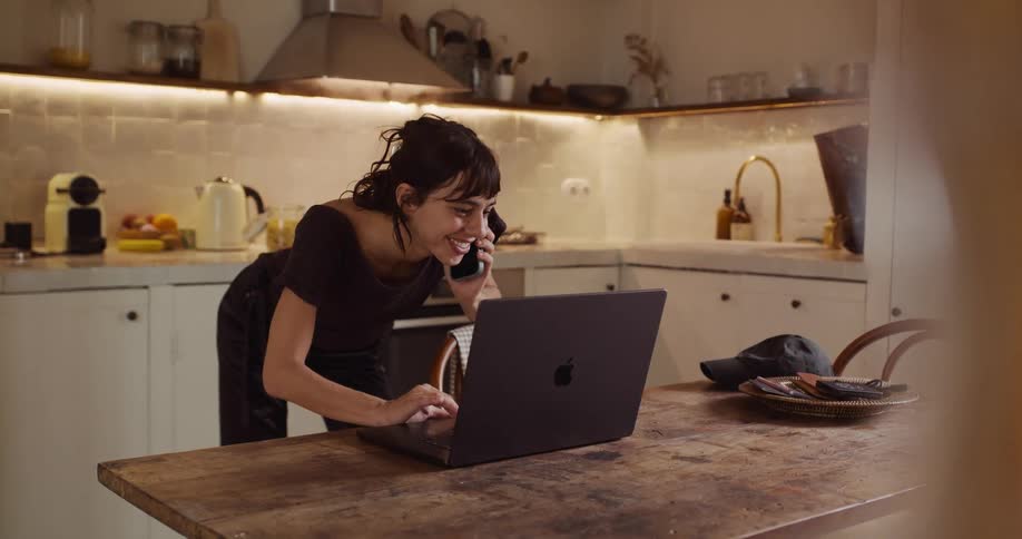 A young woman working from home in her kitchen smiles with excitement while talking on the phone and using her laptop