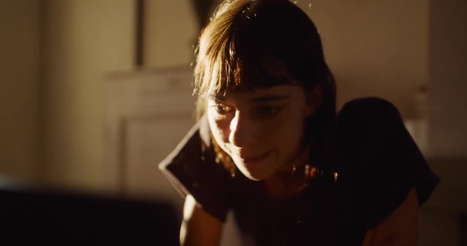 A young woman smiles contentedly while browsing on her laptop in a dimly lit room with warm, natural light