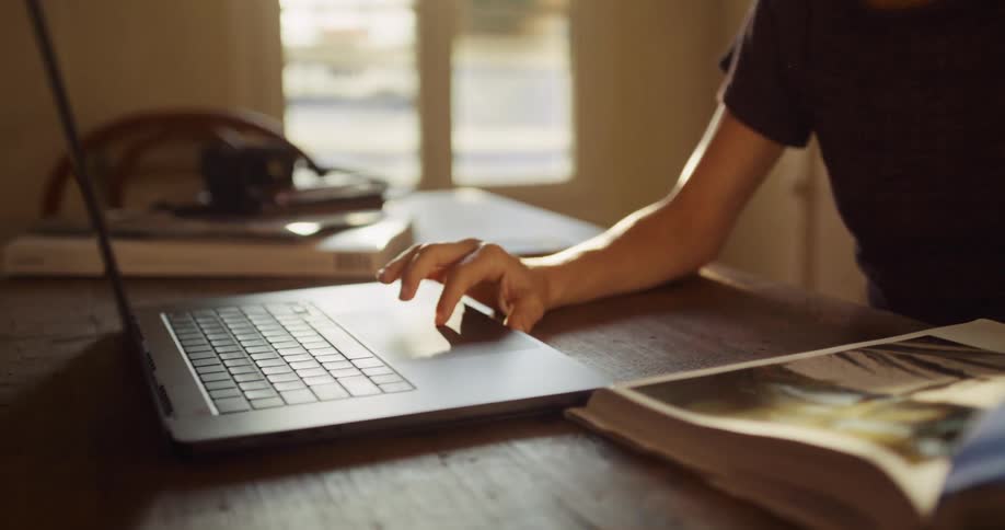 A young woman sits at a wooden table in a sunlit room, studying a photo book and using her laptop