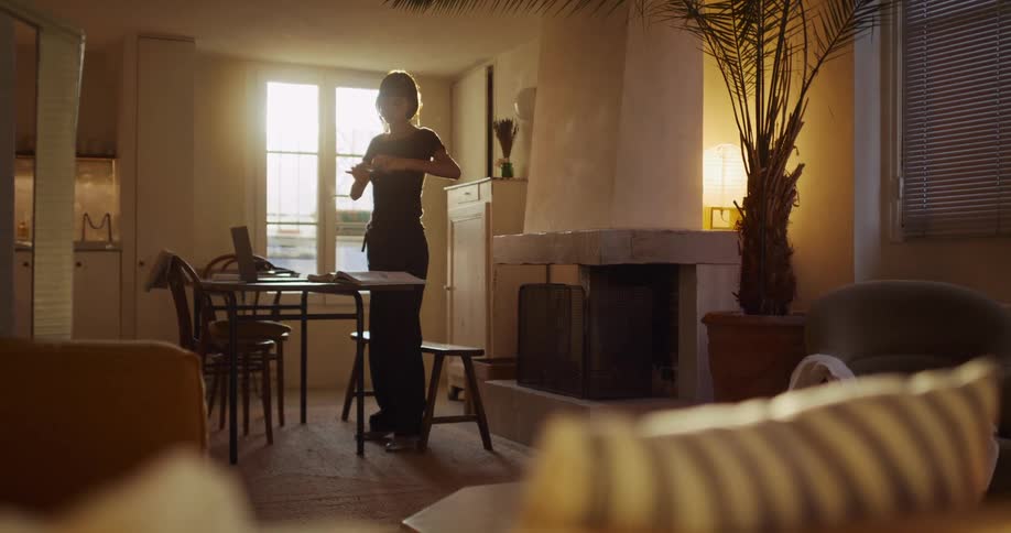 Young woman checks her phone before sitting down to work on her laptop in a cozy, sun-drenched city apartment
