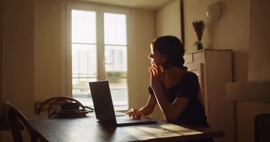 A thoughtful woman sits at a wooden table by a window, using her laptop in the warm afternoon sunlight