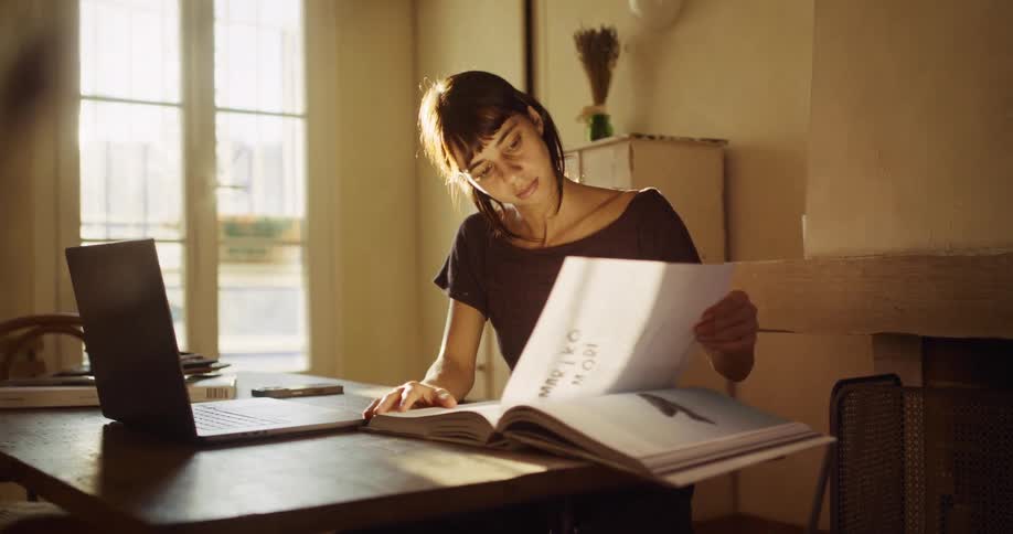 A young woman works from home, researching on her laptop and in a book at a sun-drenched desk