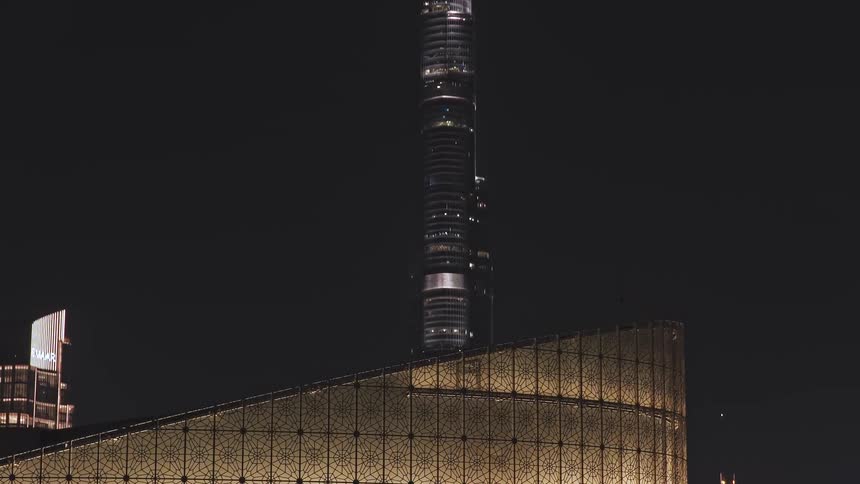 A stunning drone shot reveals the iconic Burj Khalifa and the sparkling Dubai cityscape at night, rising behind a modern architectural masterpiece