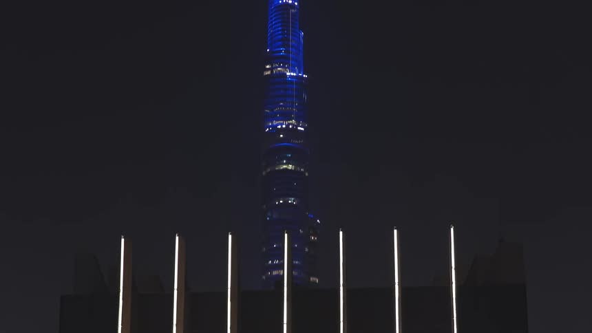 Aerial drone shot reveals the iconic blue-lit Burj Khalifa behind a modern skyscraper, zooming out to show the dazzling Dubai city lights at night