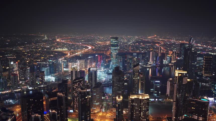 A breathtaking slow push-in aerial shot reveals the dazzling Dubai cityscape at night, with iconic skyscrapers reflecting in the water and highways aglow with traffic
