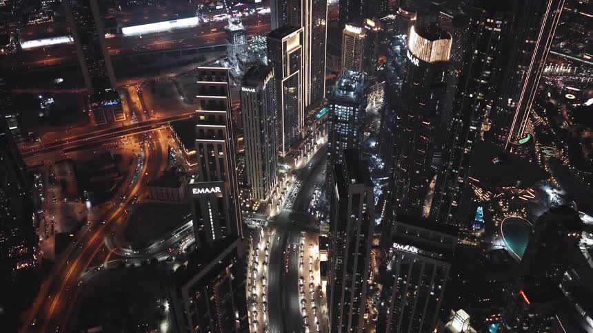 A dramatic high-angle aerial view tilts down over the brightly illuminated skyscrapers and bustling streets of Downtown Dubai at night