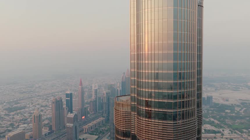 An aerial view pulls back from a close-up of the sun's reflection on the Burj Khalifa to reveal the hazy Dubai cityscape at sunset