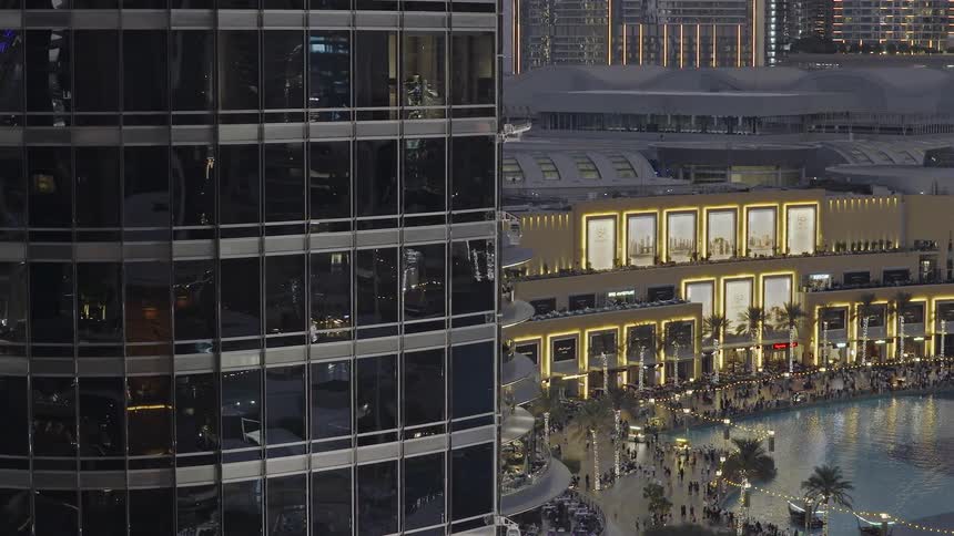 Slow zoom out from a reflective skyscraper reveals the bustling Dubai Mall and promenade at dusk