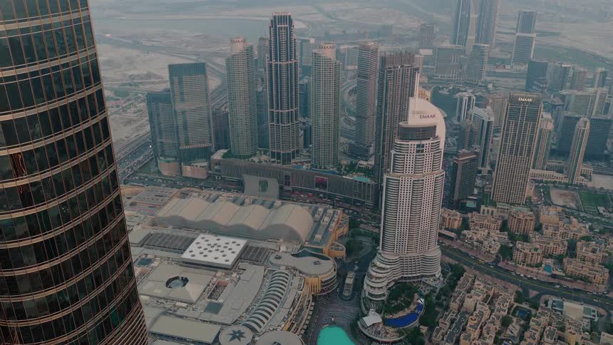 An aerial view pulls back from the reflective glass of the Burj Khalifa at sunset to reveal the stunning Dubai cityscape, including the Dubai Mall and the iconic fountain lake
