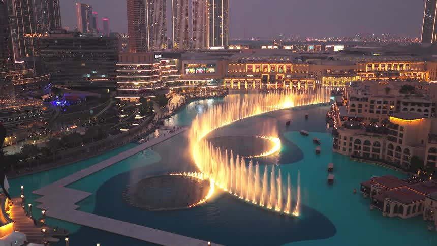 A spectacular aerial view of the Dubai Fountain show at night, with illuminated water jets dancing in front of the city's glittering skyline and the UAE flag