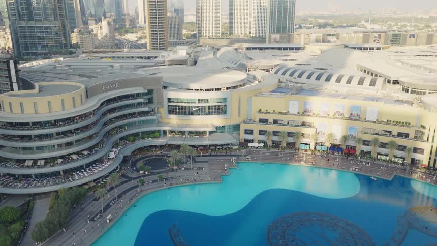 A slow aerial pullback reveals the massive Dubai Mall, the Dubai Fountain, and the surrounding city skyline