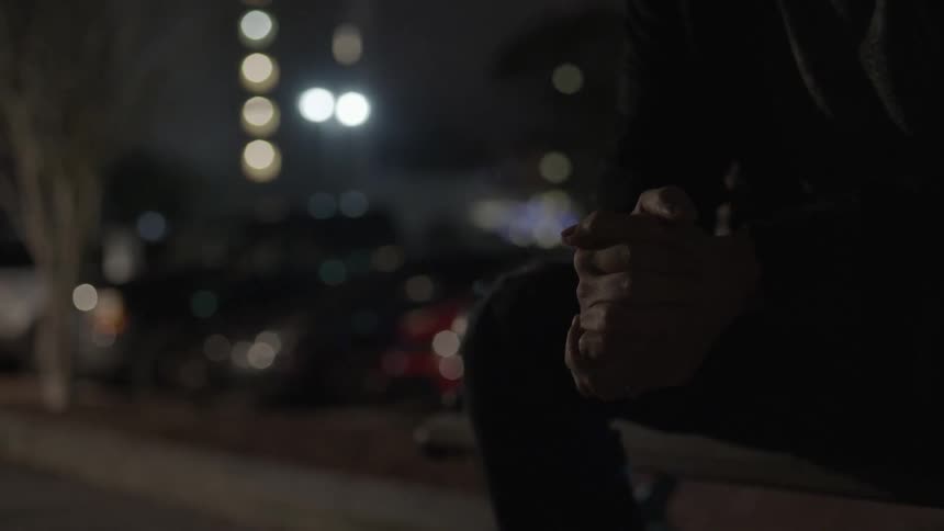A man clasps his hands together while sitting in a city setting at night