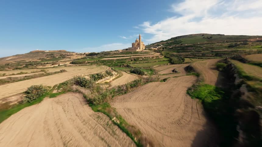 An FPV drone flies at high speed over the terraced fields and rolling green hills towards the magnificent Basilica of Ta' Pinu in Gozo, Malta