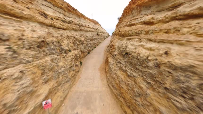 An FPV drone speeds over the turquoise ocean before flying through a narrow canyon carved into the rocky coastline