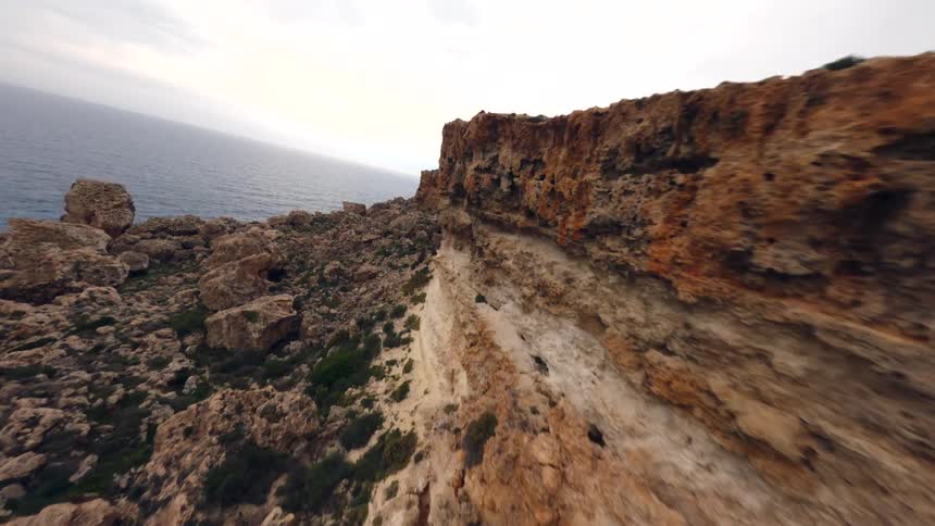 FPV drone footage captures a dynamic flight along a rugged, rocky cliffside bordering the tranquil ocean under a cloudy sky